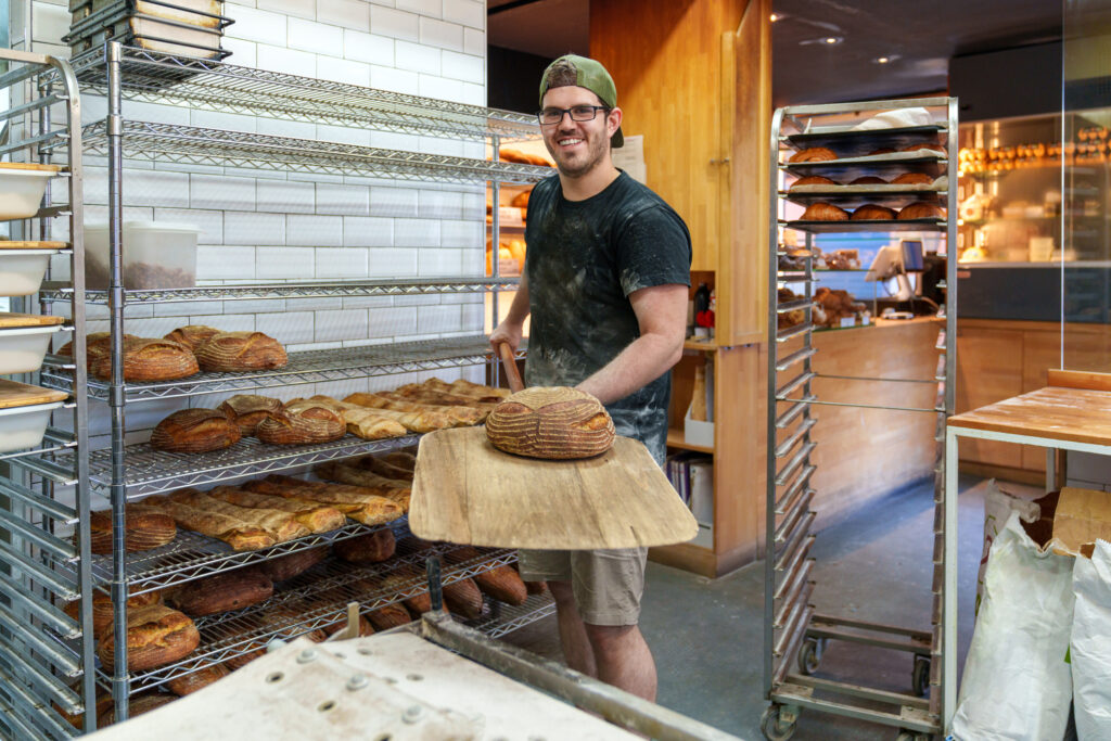 Un joyeux boulanger dans une boulangerie animée, armé d'une pelle à pain, se tient près d'un chariot rempli de pains fraîchement cuits, prêts pour le four.