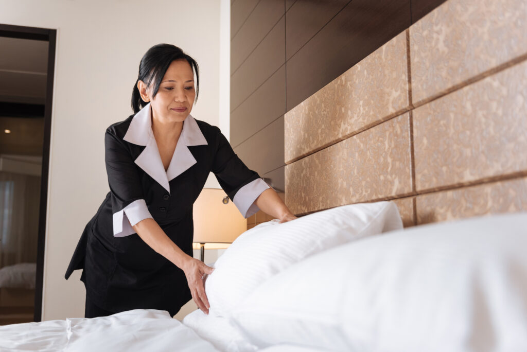 Part of my duties. Pleasant delighted Asian hotel maid standing over the bed and putting a pillow while making it